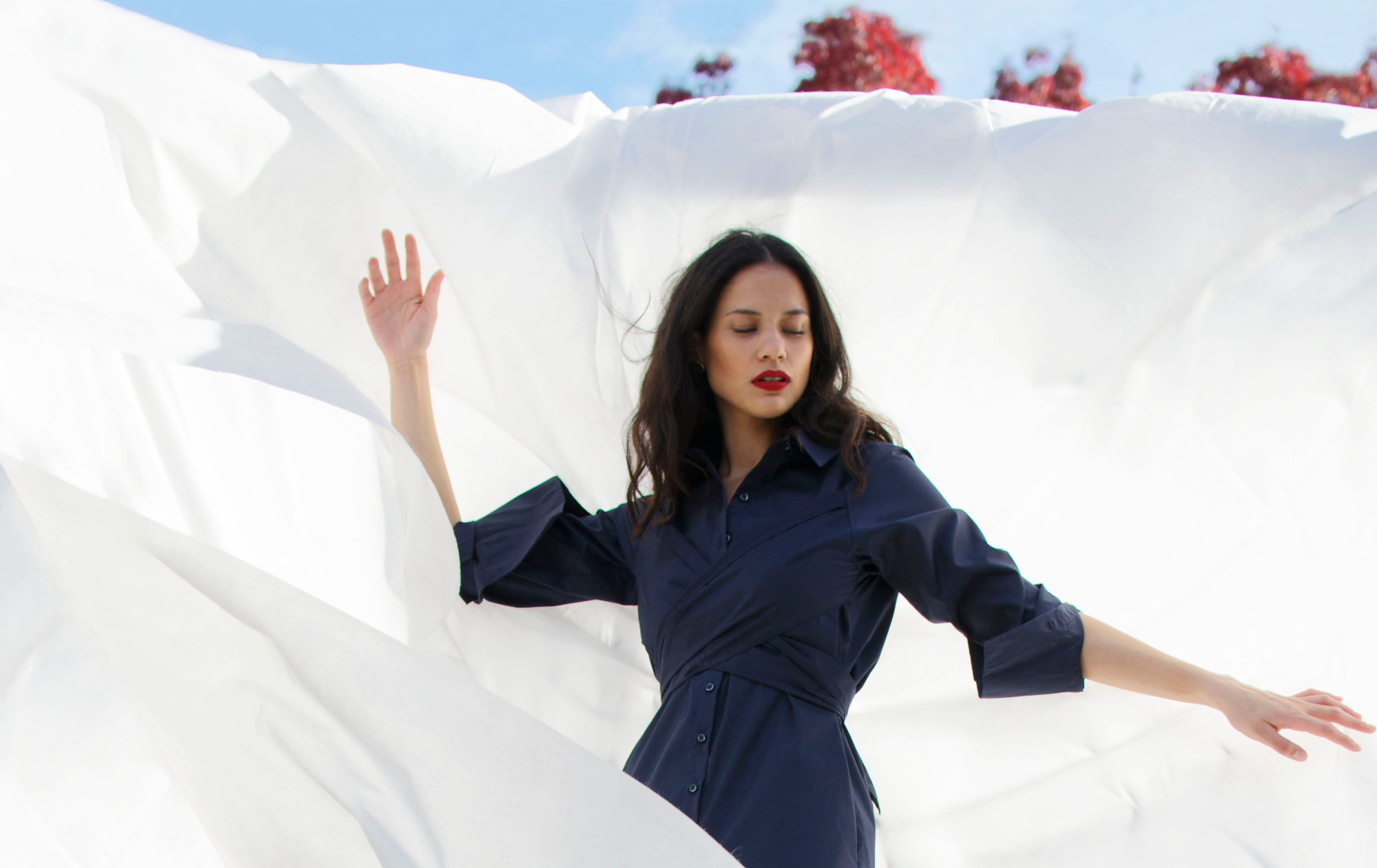 Woman in a blue dress standing amidst flowing white fabric with a clear sky and trees in the background